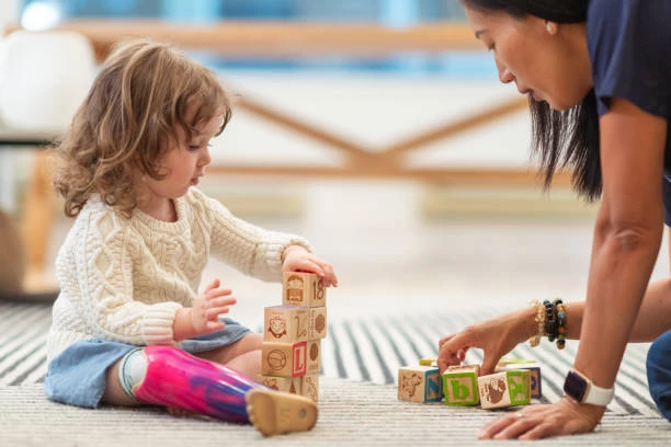 A therapist moves word blocks around next to a toddler stacking another pile of blocks.